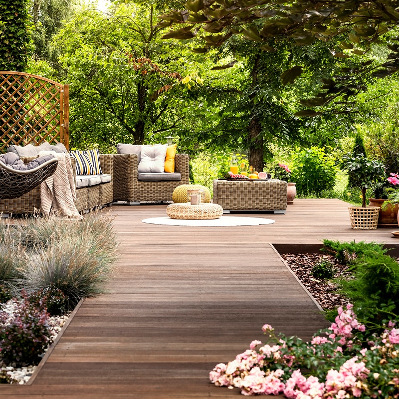 Spacieuse terrasse en bois sombre avec salon de jardin en osier, coussins gris et jaunes, entourée de plantes luxuriantes et fleurs.