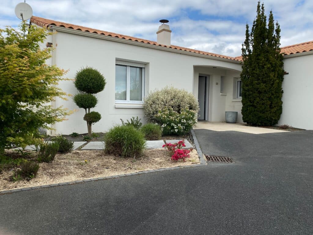 Maison moderne blanche avec toit tuile rouge, jardin paysager varié (érable, topiaire, fleurs) et allée goudronnée. Numéro 2 sur la porte d'entrée.