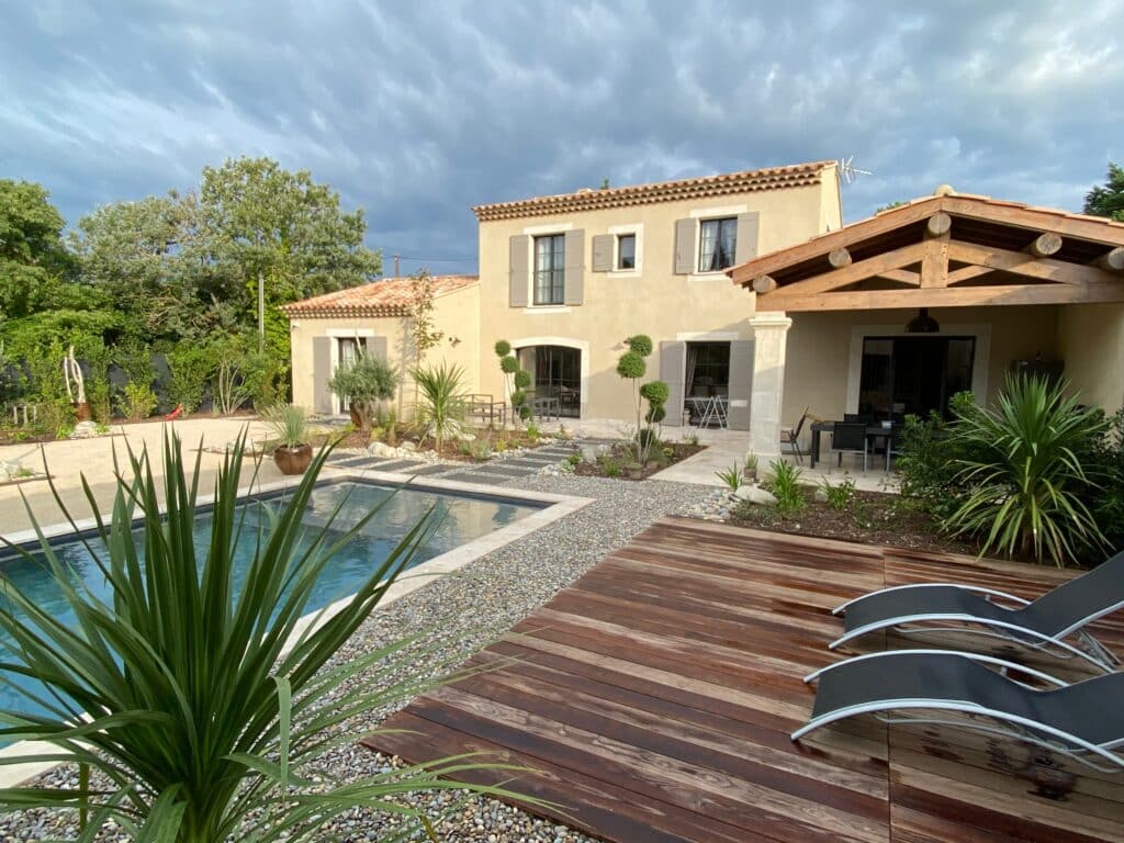 Maison moderne avec piscine, terrasse en bois, transats et jardin méditerranéen sous ciel nuageux.